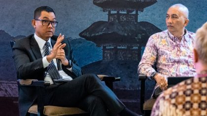 Picture of two men speaking at an event in front of a blue backdrop featuring traditional Balinese architecture. One man wears a suit and tie, the other a batik shirt.
