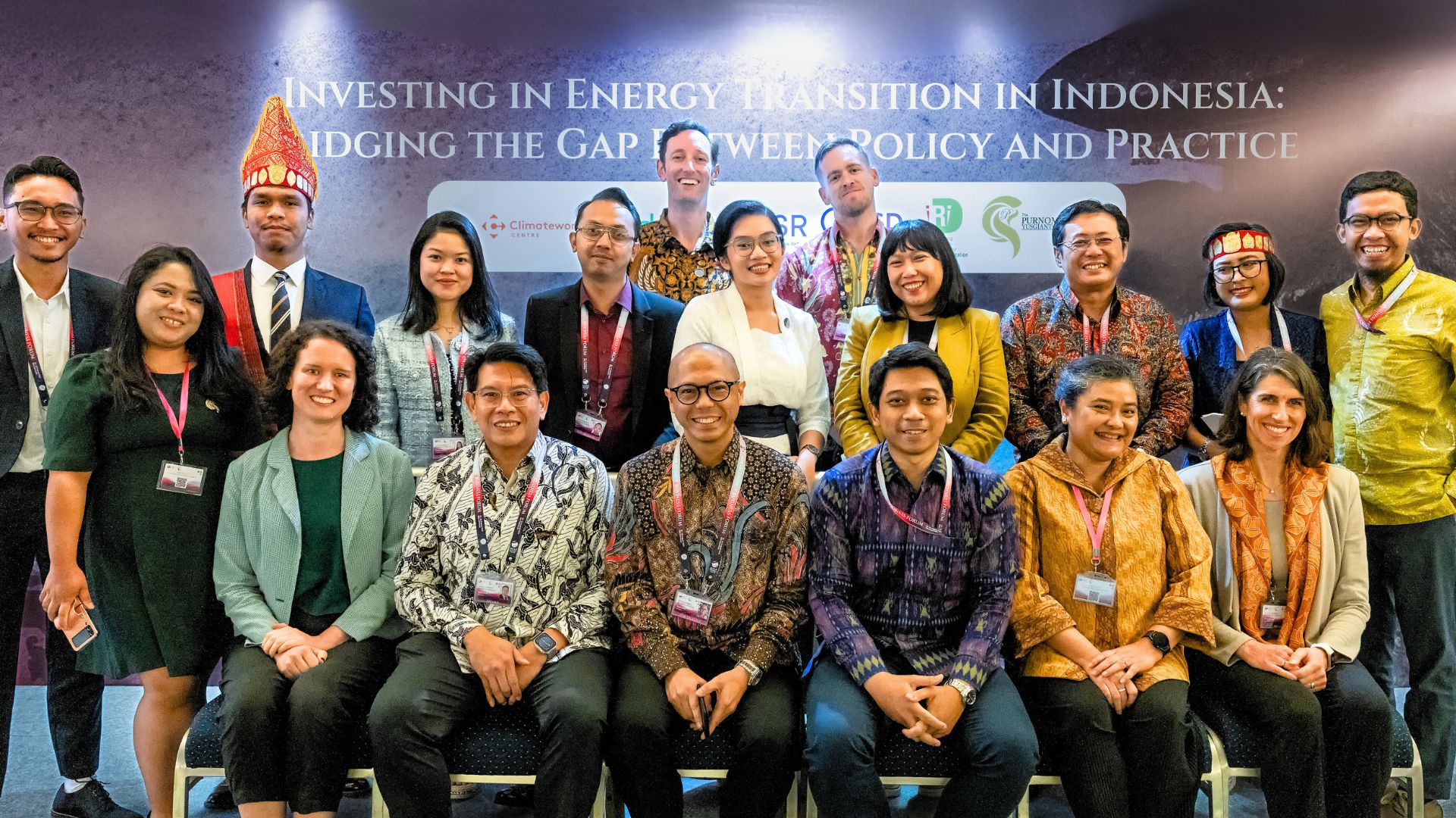 A group photograph of men and women in formal western and Indonesian attire smiling at the camera. Behind them sits a backdrop reading "Investing in Energy Transition in Indonesia: Bridging the gap between Policy and Practice"